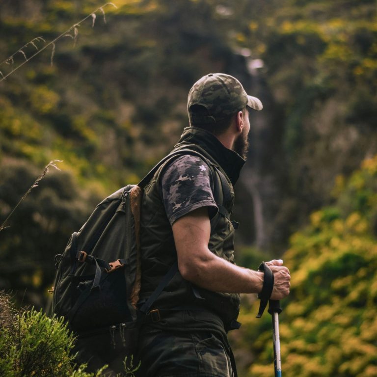 A missionary nomad Man in camouflage stands on a trail, looking at a waterfall surrounded by greenery.